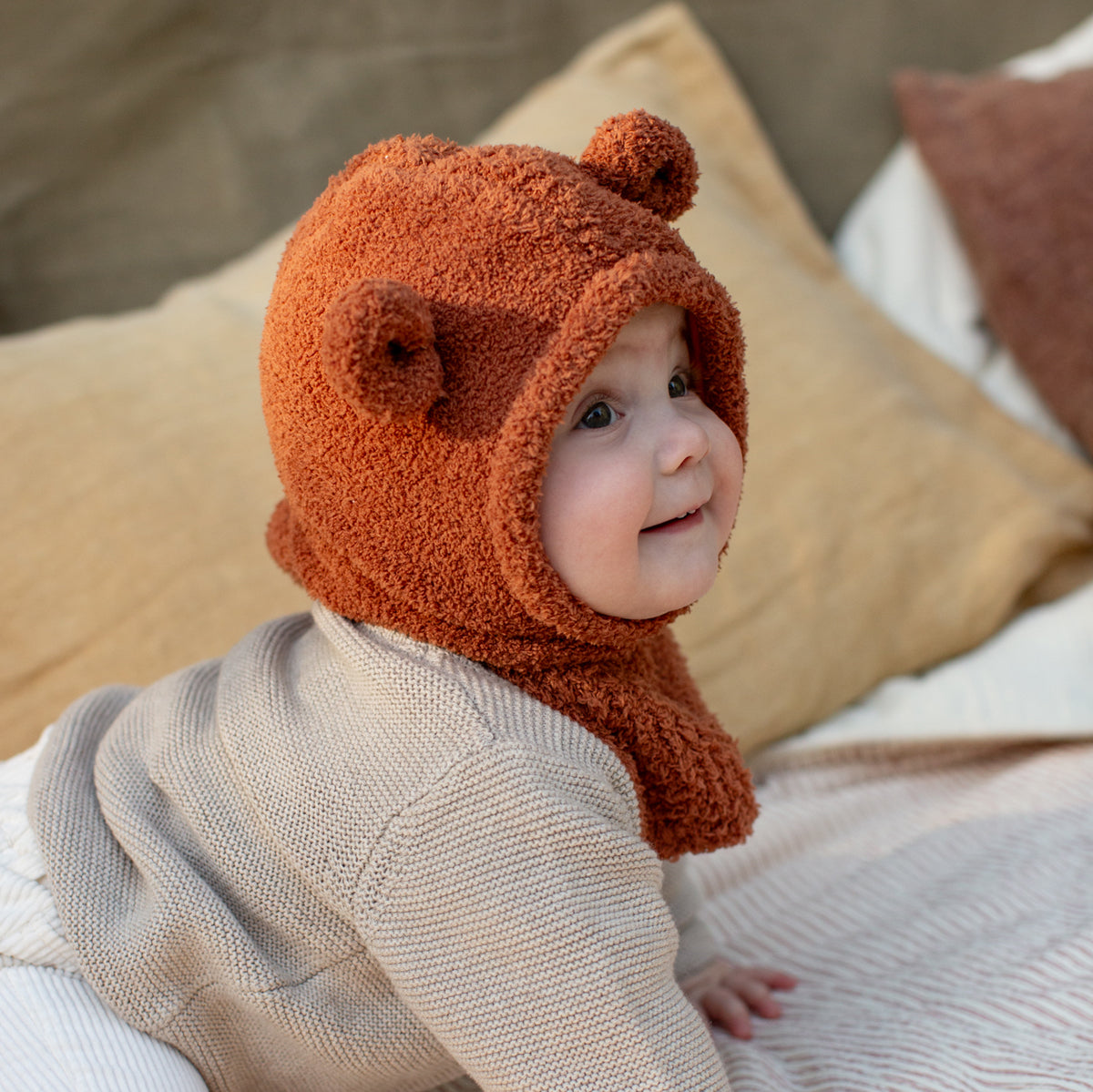 Baby wearing a brown teddy bear hat on a bed with pillows