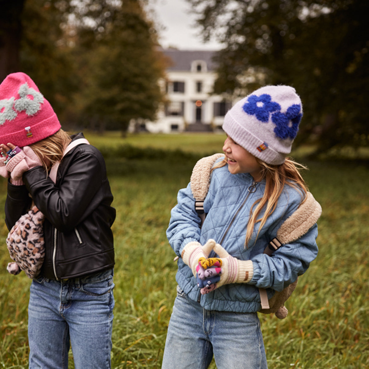 Two young girls standing in a grassy yard outdoors. The girl on the right wears a light blue quilted jacket with a beige sherpa lining, a white knit beanie with blue flowers, and cream knit gloves while holding colorful pom-poms.