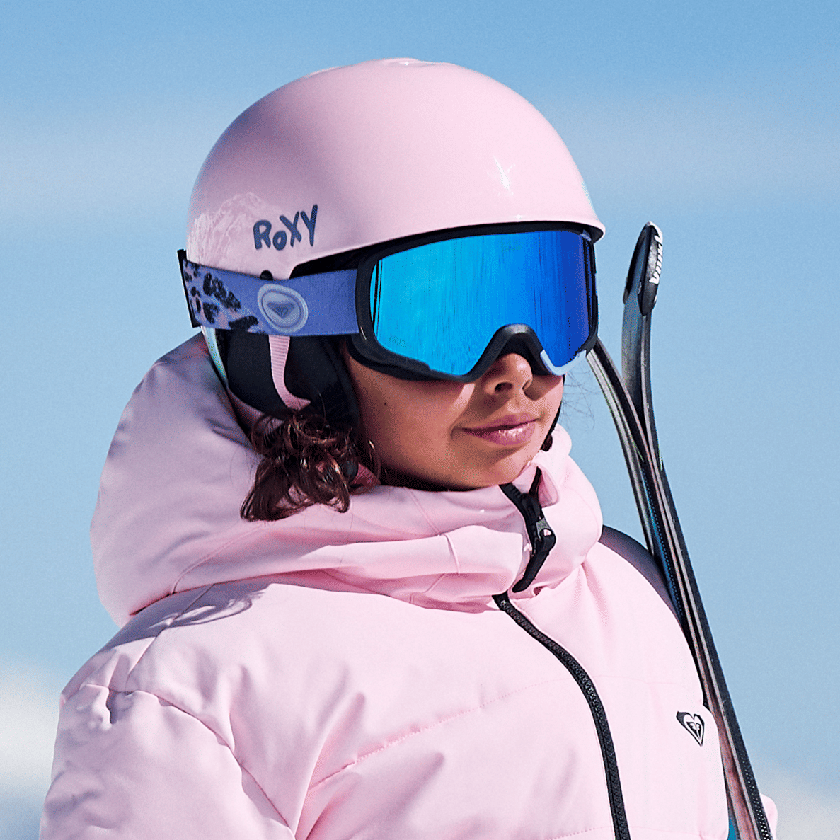 A close-up of a person wearing a light pink Roxy ski helmet, blue mirrored ski goggles, and a matching pink ski jacket, with skis visible over the shoulder against a clear blue sky.