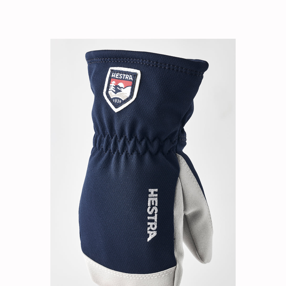 A single Hestra winter glove in navy blue and light gray, shown against a plain light gray background. The glove features a navy blue fabric body, a light gray palm, and a white Hestra shield logo patch on the cuff.