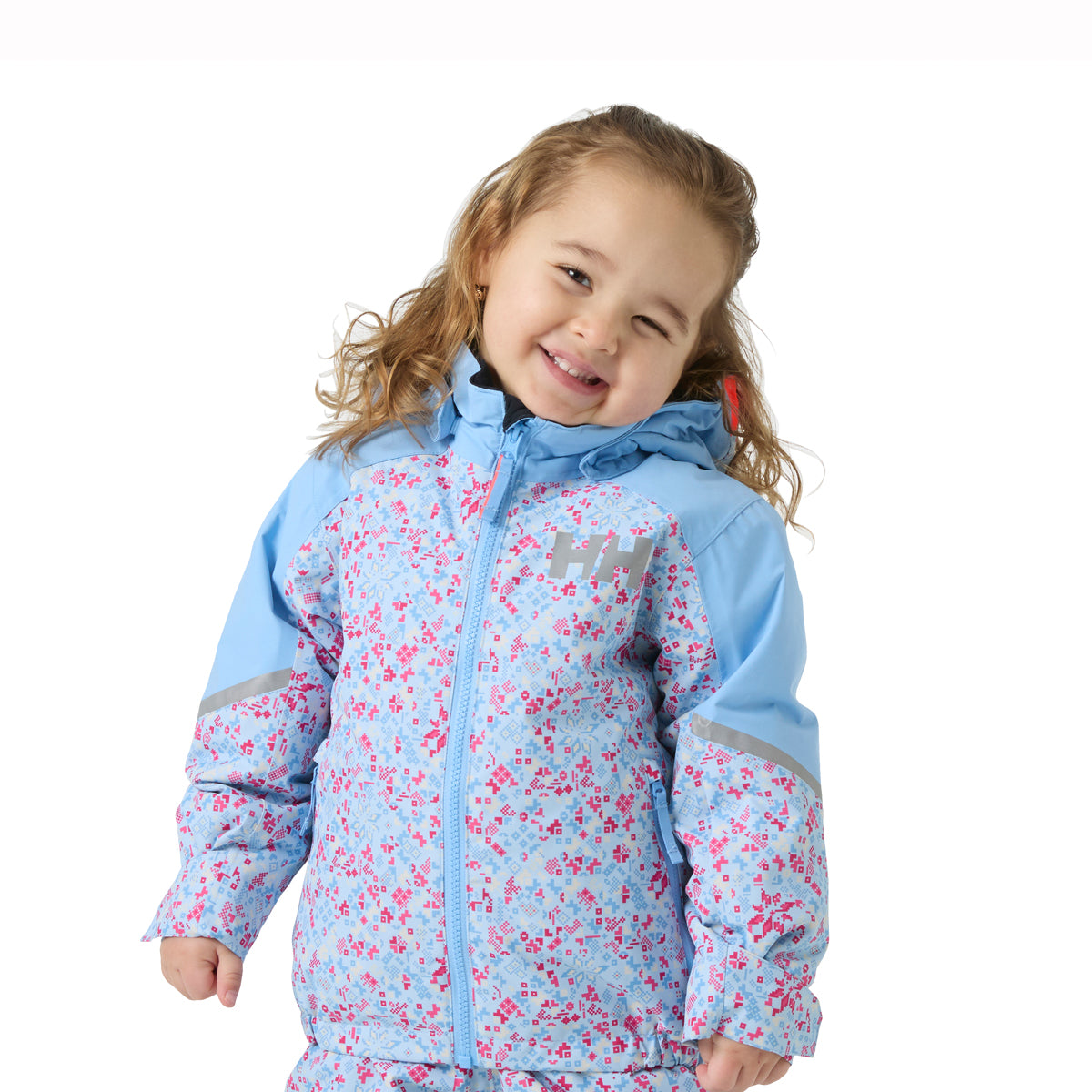 A young girl smiling and winking while wearing a light blue Helly Hansen ski jacket with a pink floral pattern, set against a plain white background.