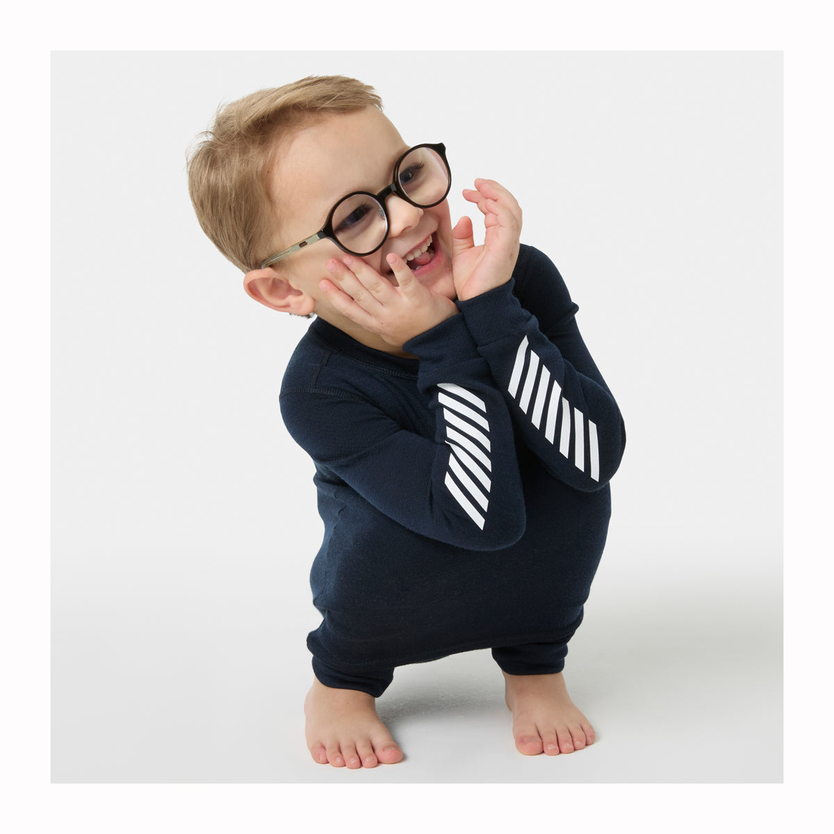 A baby wearing a navy blue long-sleeve jumpsuit with white diagonal stripes on the sleeves, styled with round black glasses, posing on a light gray studio background.