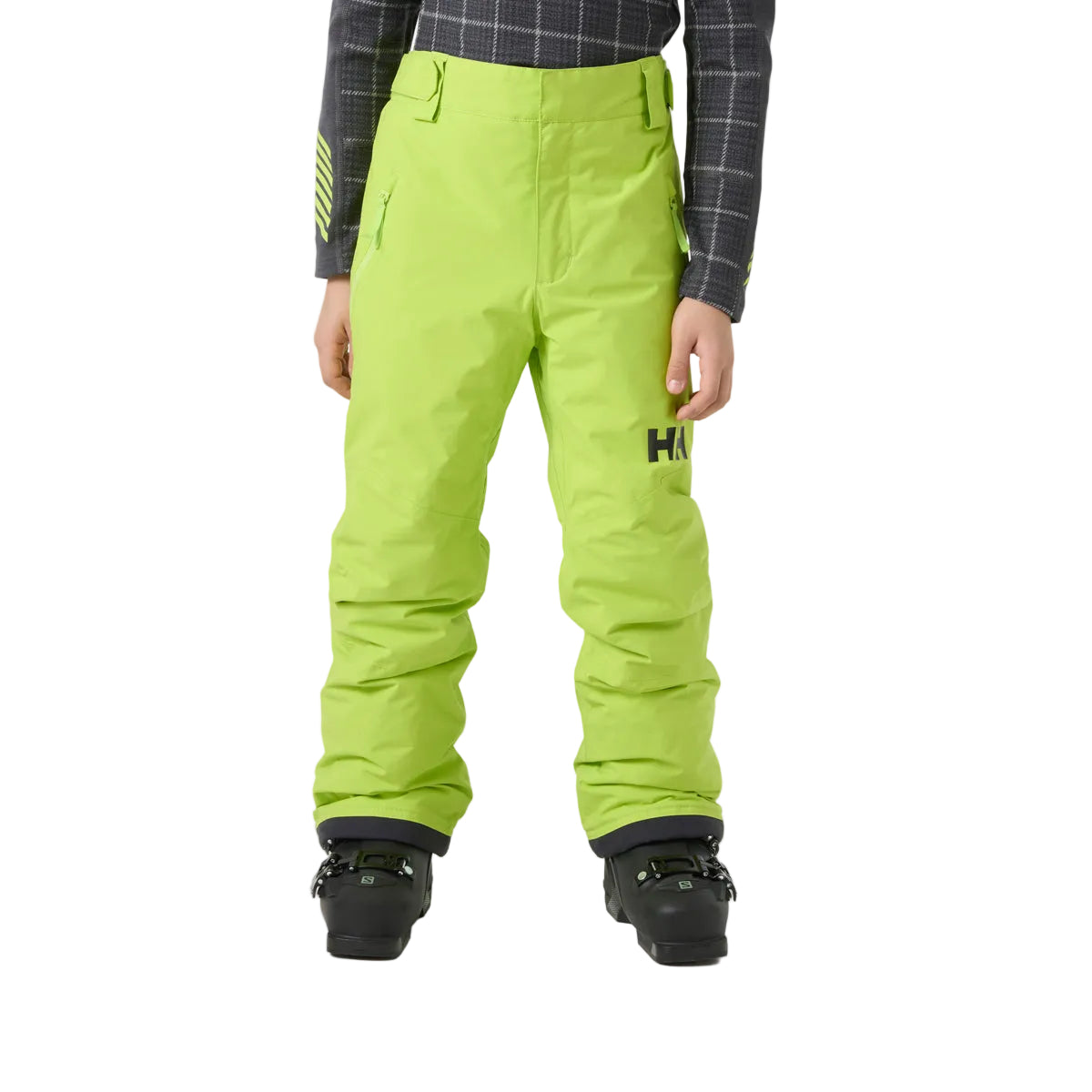 A child wearing bright lime green Helly Hansen snow pants, paired with a dark gray plaid shirt and black ski boots, shown from the waist down against a light gray studio background.