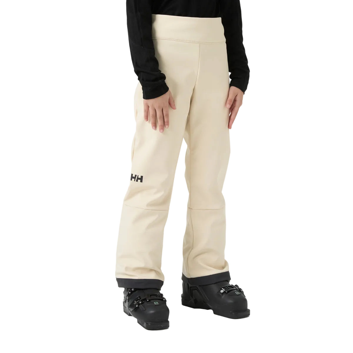A person wearing cream-colored Helly Hansen snow pants with a black long-sleeve top and black ski boots, shown from the waist down on a white studio background.