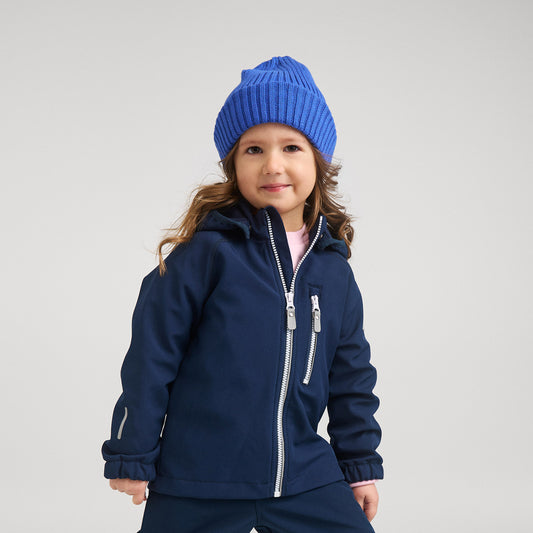 A young child wearing a navy blue hooded jacket with silver zippers, a matching navy blue beanie, and navy pants, photographed against a light gray studio background.