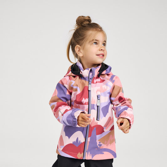 A young girl with her hair in a bun is wearing a hooded ski jacket with a pink, purple, and red abstract landscape pattern, standing against a light gray studio background.