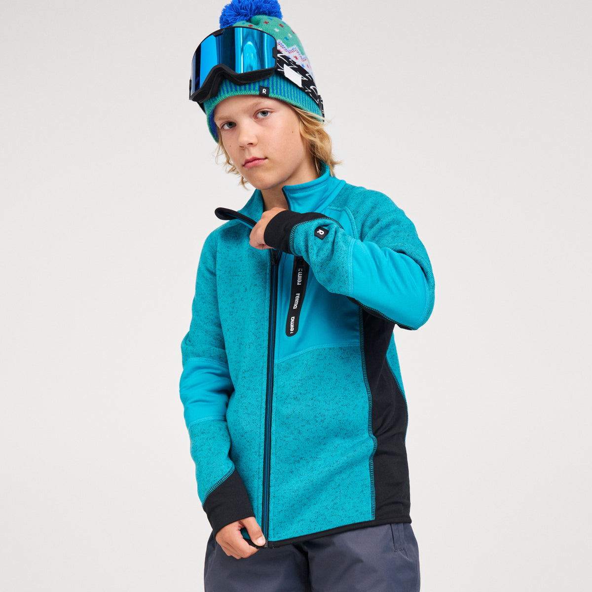 A child wearing a bright turquoise and black ski fleece jacket, a matching knit beanie with a blue pom-pom, and ski goggles, posed against a light gray studio background.