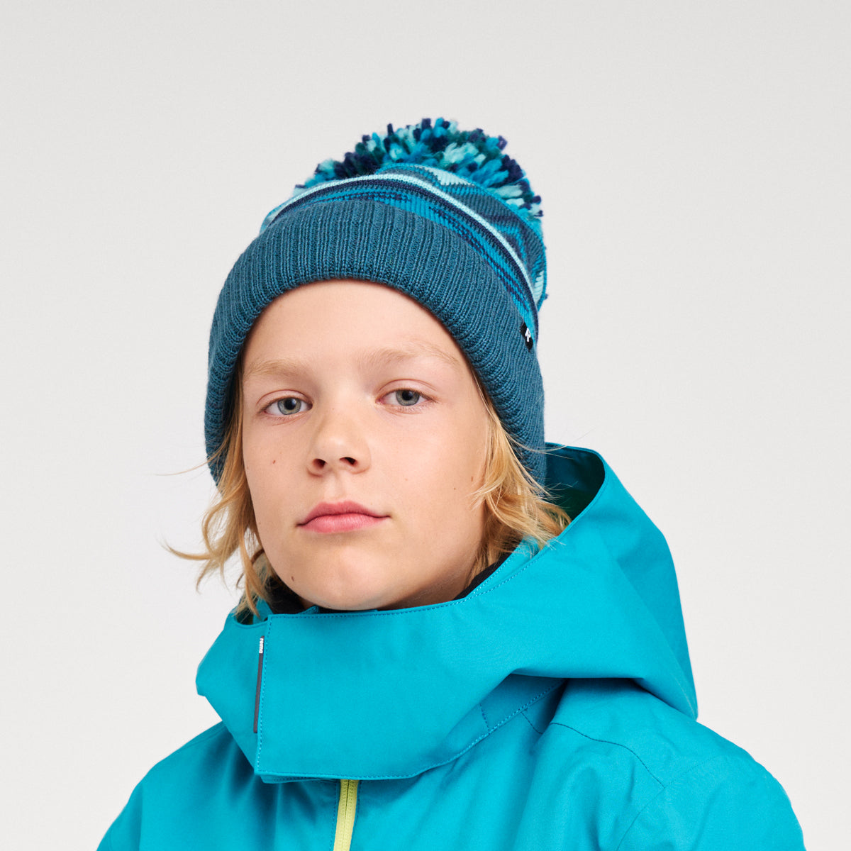 A child wearing a teal blue knit beanie with a blue and teal pom-pom and a bright turquoise hooded jacket, photographed against a light gray studio background.