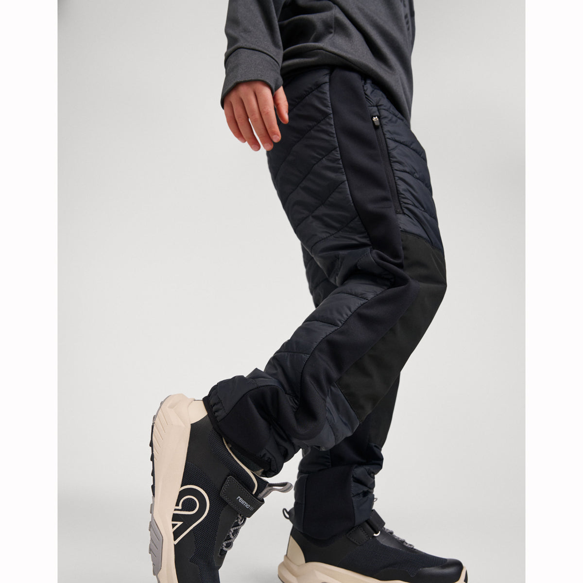 A person wearing black quilted ski pants with zippered knee panels and black athletic boots, shown from the waist down against a light gray studio background.