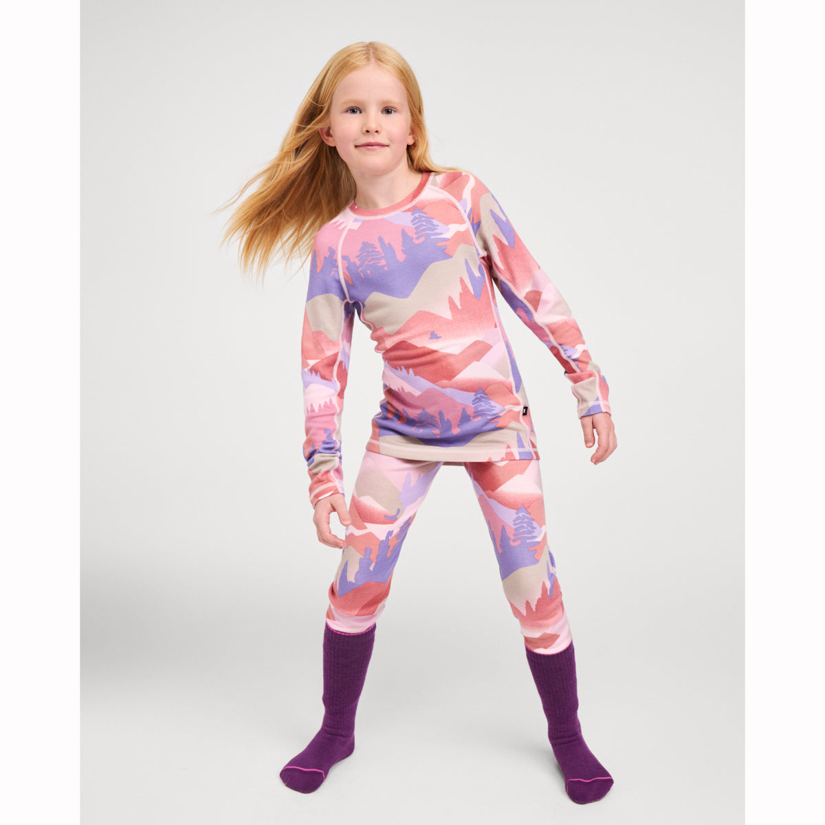A young girl modeling a matching two-piece long-sleeve pajama set with a pink, purple, and beige mountain landscape print, paired with solid purple socks, against a light gray studio background.