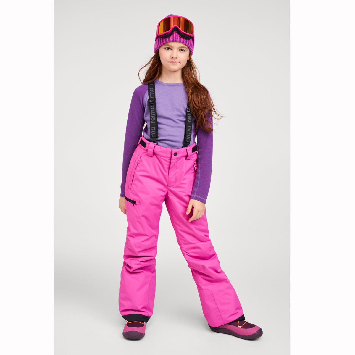 A young girl wearing a pink and purple ski outfit, including bright pink snow pants with black suspenders, a purple long-sleeve shirt, a pink beanie, and ski goggles, standing against a light gray studio background.