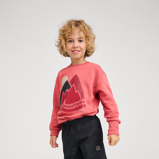A young child with curly blonde hair wearing a coral pink crewneck sweatshirt with a mountain graphic and black pants, standing against a light gray studio background.