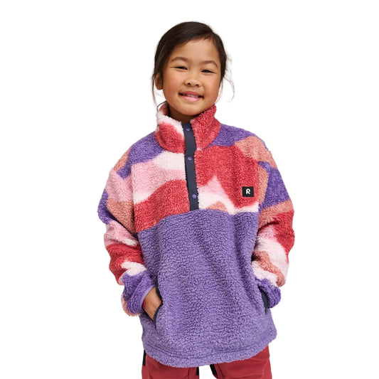 A young girl smiling while wearing a colorful sherpa fleece pullover jacket with a purple front panel and wavy red, pink, and purple patterns on the sleeves and upper chest, set against a plain light gray studio background.