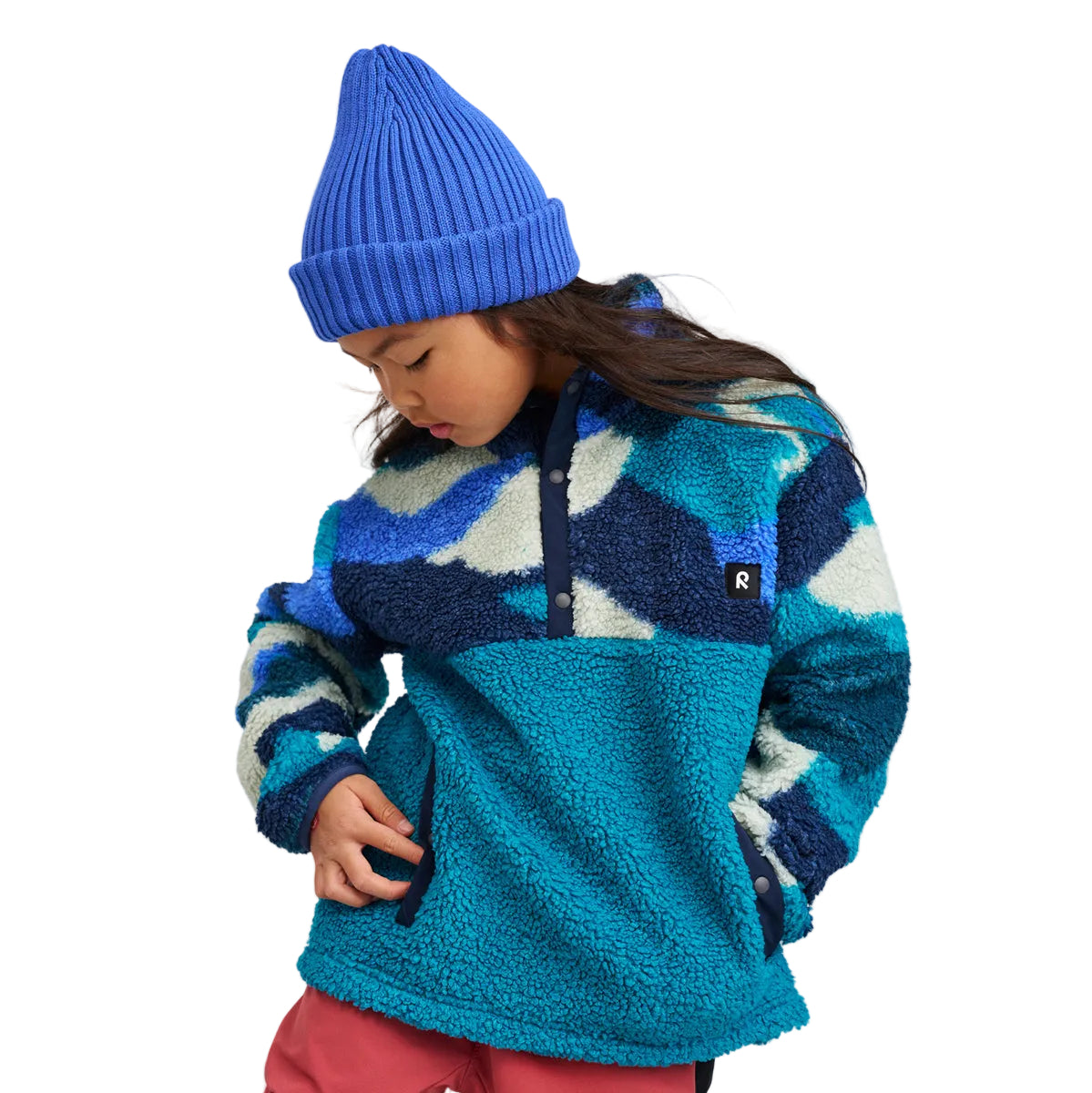 A child wearing a blue ribbed knit beanie and a colorful sherpa fleece pullover jacket with a blue and white geometric pattern, looking down against a light gray background.