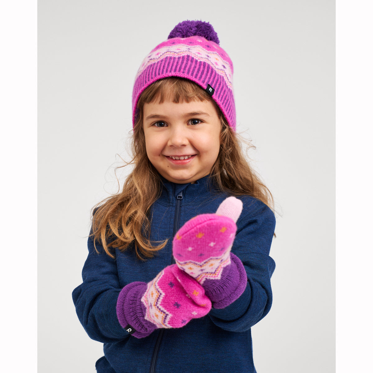 A young girl smiling and wearing a pink and purple knit beanie with a pom-pom, matching knit mittens, and a navy blue fleece jacket against a light gray studio background.