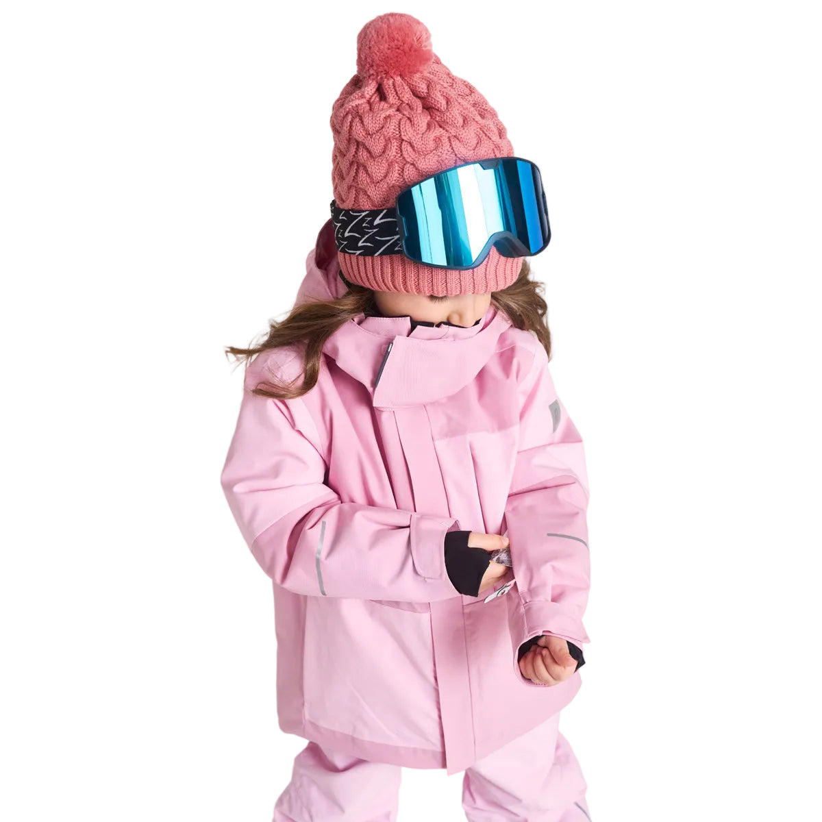 A child wearing a light pink ski jacket and matching pants, along with a pink knit beanie and blue mirrored ski goggles, standing against a light gray studio background.