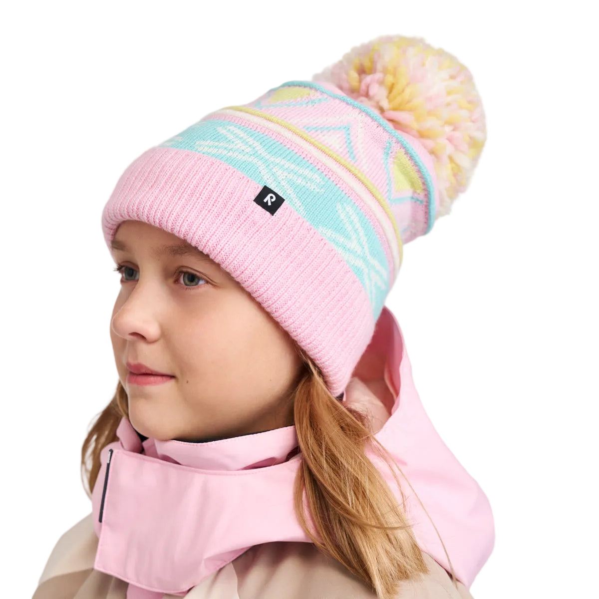A child wearing a pink knit beanie with a multicolor geometric pattern and a fluffy pom-pom, paired with a light pink jacket, photographed against a plain light gray background.