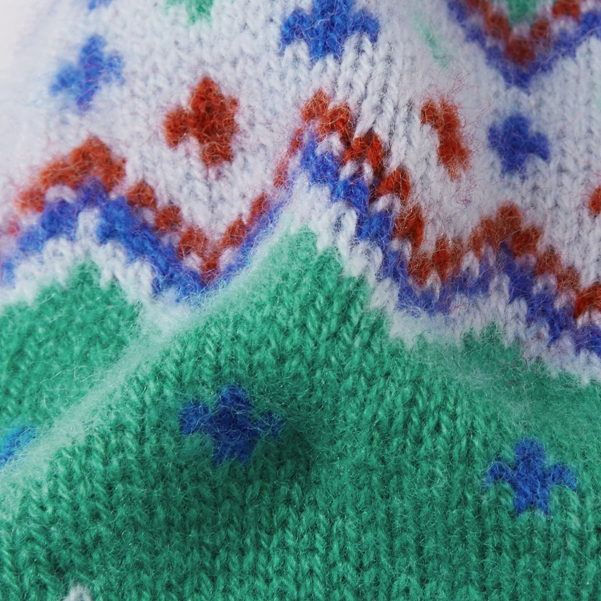 Close-up of a knitted textile with a multicolor pattern, featuring a green lower section, a white upper section with red and blue motifs, and blue star-like accents.