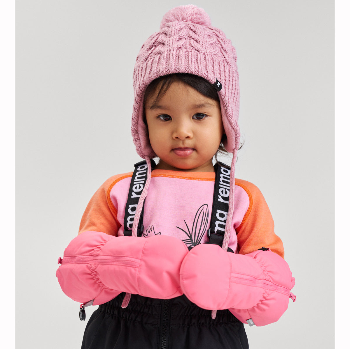 Child wearing a pink knit hat, pink mittens, and an orange and black jacket with visible branding.