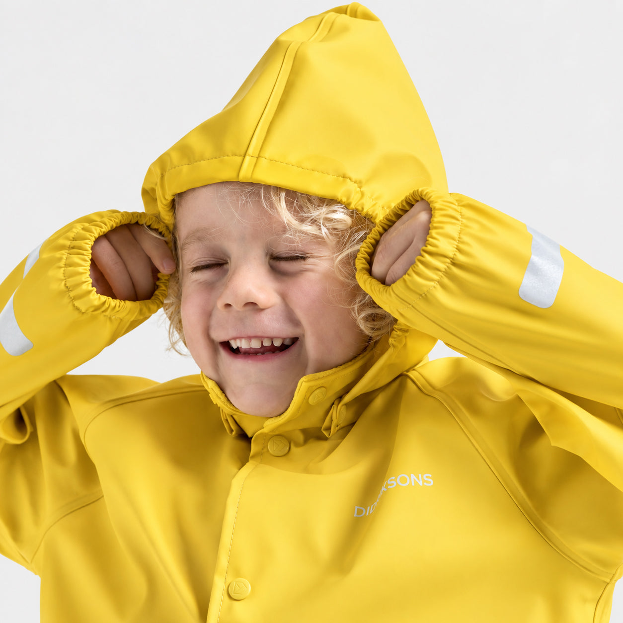 A smiling young child wearing a bright yellow hooded rain jacket with reflective white stripes on the cuffs, posing with hands in the hood against a light gray background.