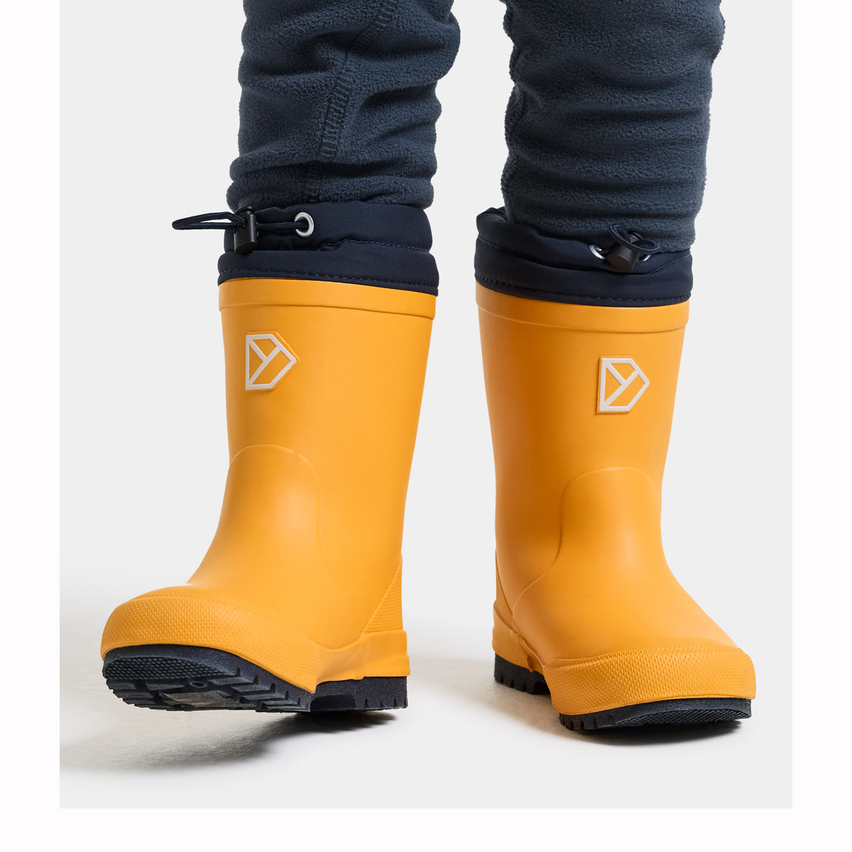A pair of bright yellow rubber rain boots with black soles and black cuffs, shown on a child's feet wearing dark gray fleece pants against a light gray studio background.