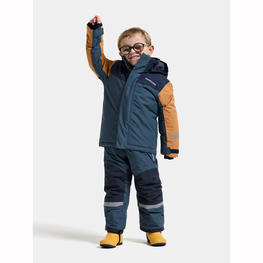 A young child wearing a navy and tan ski jacket and matching pants with yellow boots, standing against a light gray studio background.