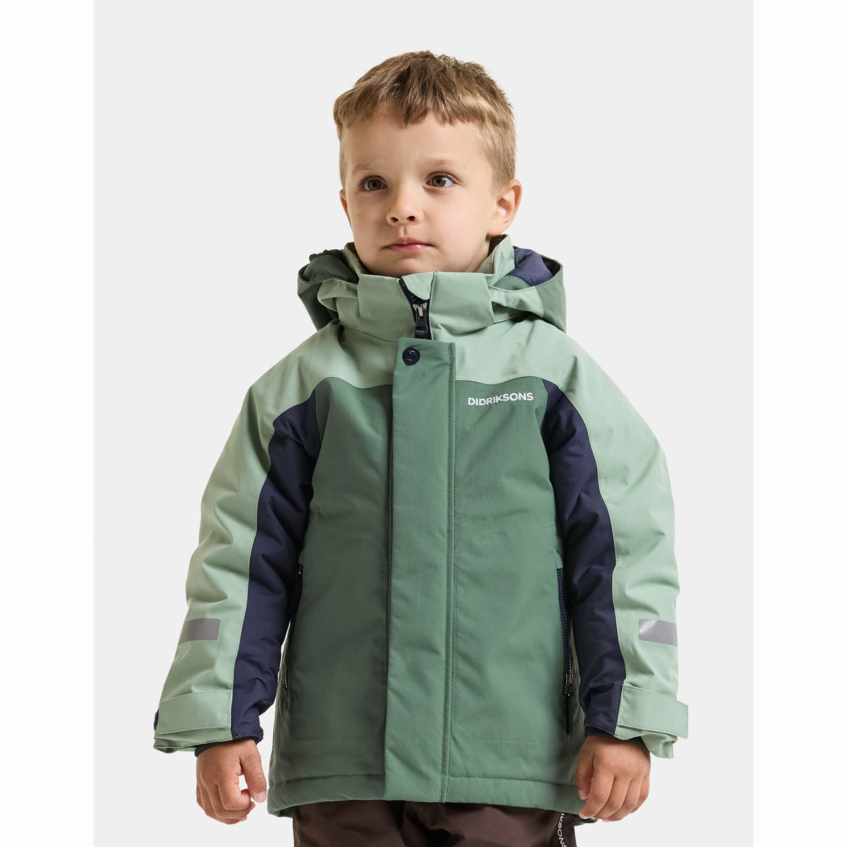 A young boy wearing a green and navy blue hooded winter jacket with the brand name 'Didriksons' printed on the chest, set against a plain light grey background.