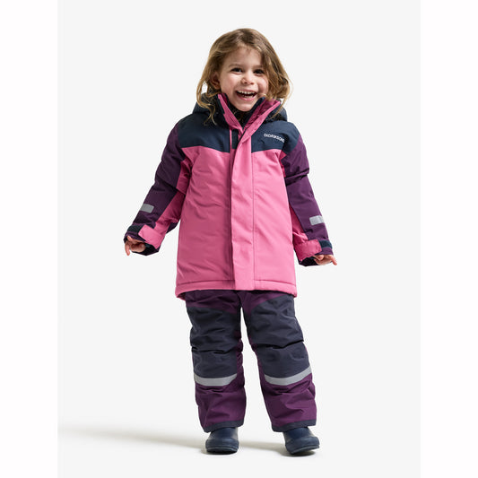 A young child wearing a pink and dark navy winter jacket and matching snow pants with reflective stripes, standing against a plain light gray studio background.