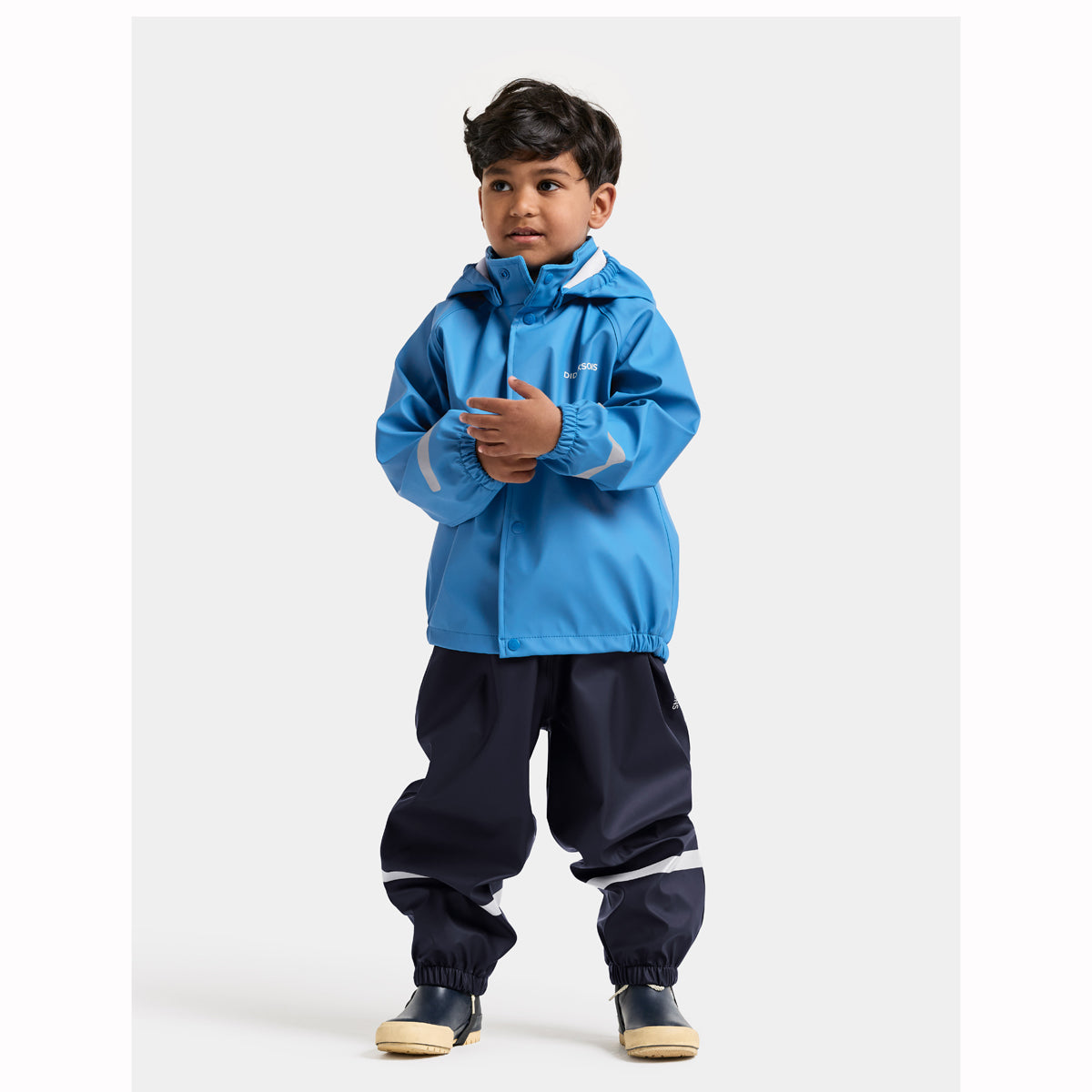 A young boy wearing a bright blue hooded rain jacket and navy blue pants with white stripes, standing against a light gray studio background.