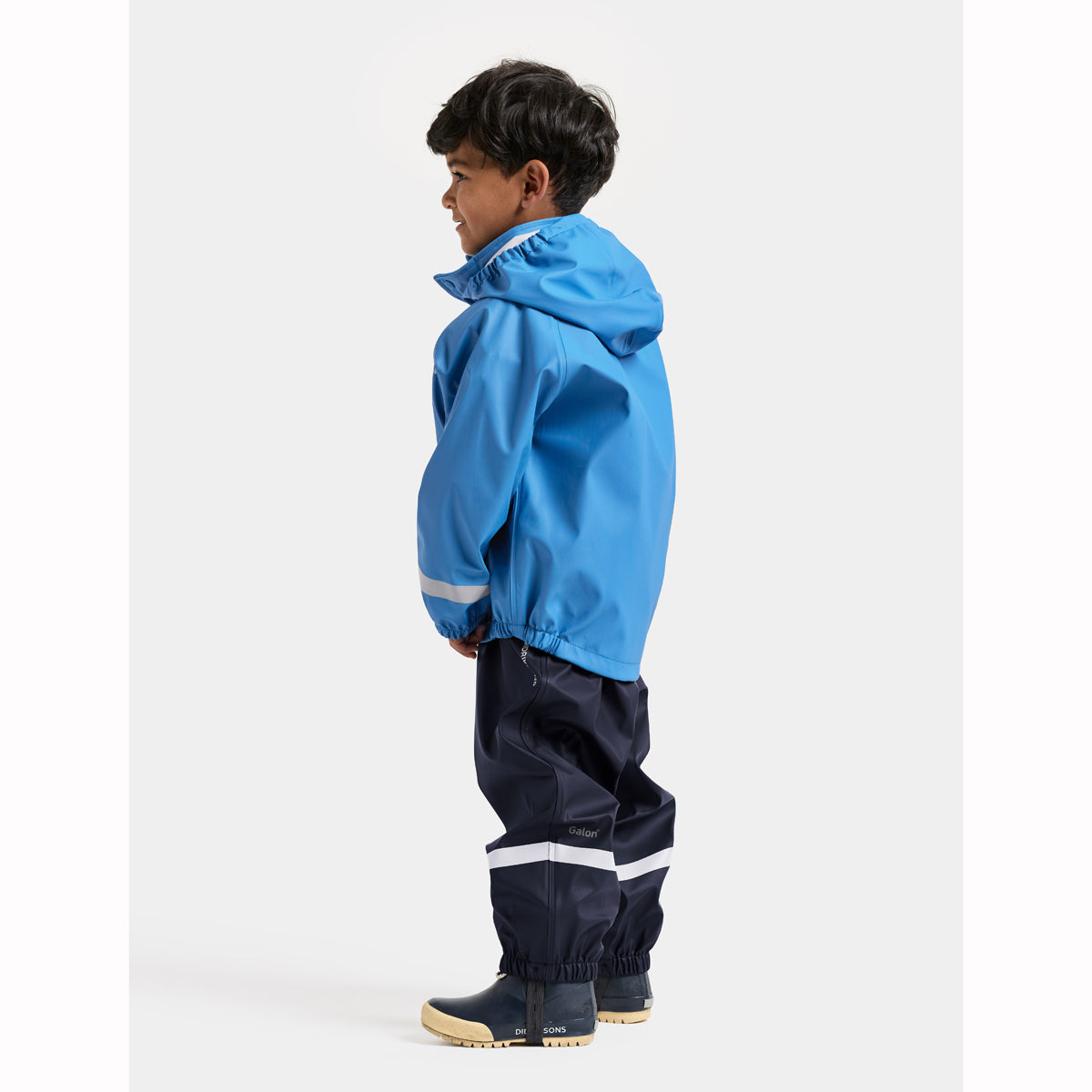 A child model shown from the side wearing a bright blue hooded rain jacket and dark navy rain pants with reflective white stripes, standing against a light gray studio background.