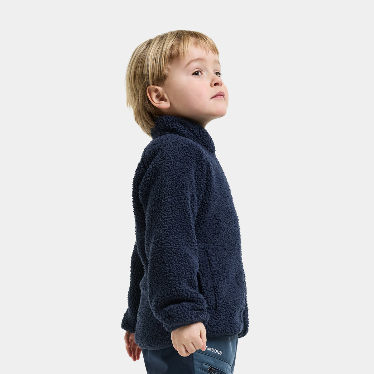 A young boy with blonde hair is shown in profile wearing a dark navy blue sherpa fleece jacket and dark pants against a light gray studio background.