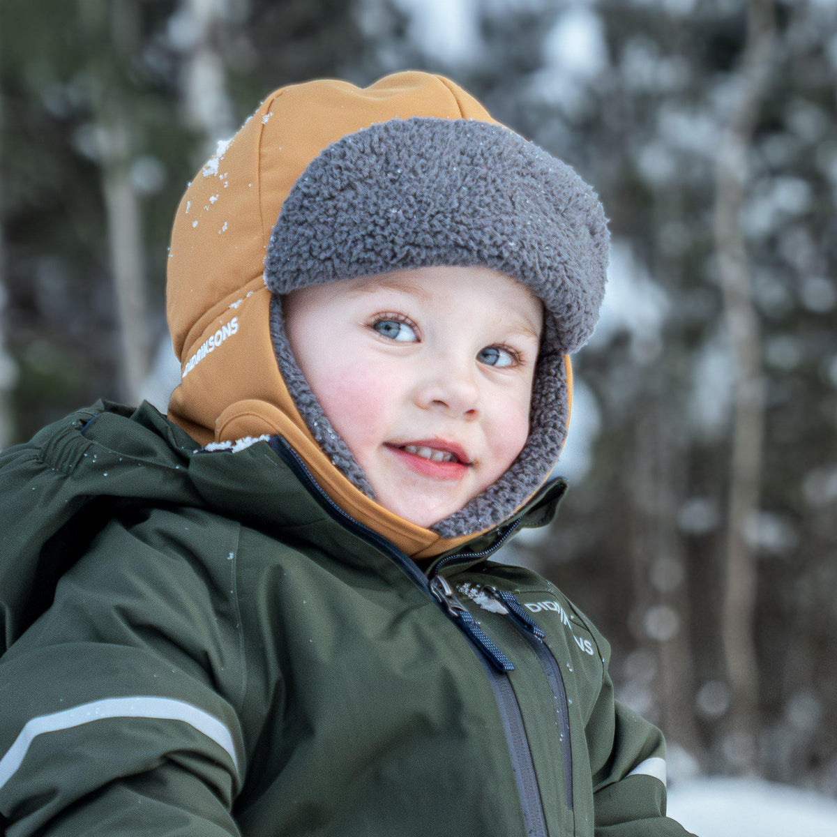 A young child wearing a dark olive green winter jacket and a tan and gray fleece winter hat, photographed outdoors in a snowy forest setting.