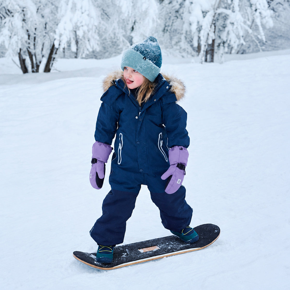 A young child wearing a navy blue snowsuit, purple gloves, and a light blue beanie stands on a snowboard in a snowy outdoor setting with snow-covered trees in the background.