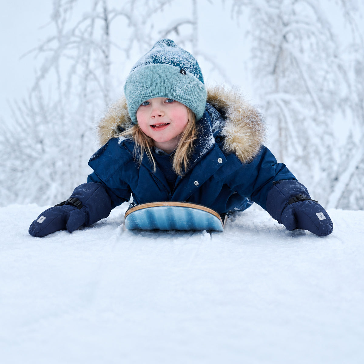 A young child wearing a navy blue winter jacket with a fur-trimmed hood, a light blue knit beanie, and navy gloves lies in the snow, smiling while holding a blue sled.