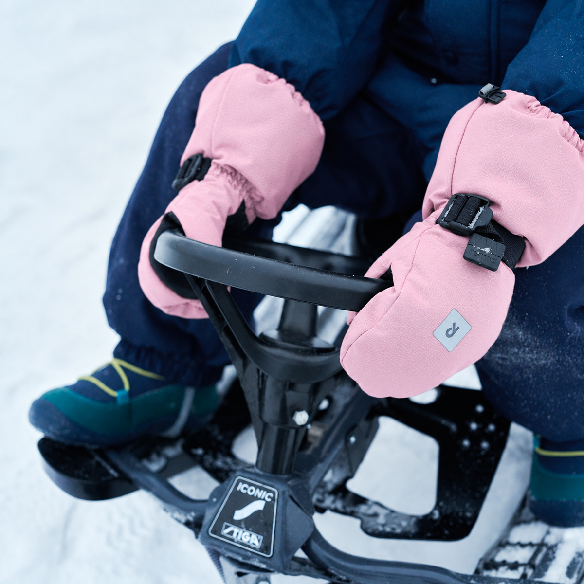 A close-up of a person wearing pink insulated winter gloves with black buckle straps, sitting on a snowmobile in the snow. The gloves have a small square logo patch on the back.