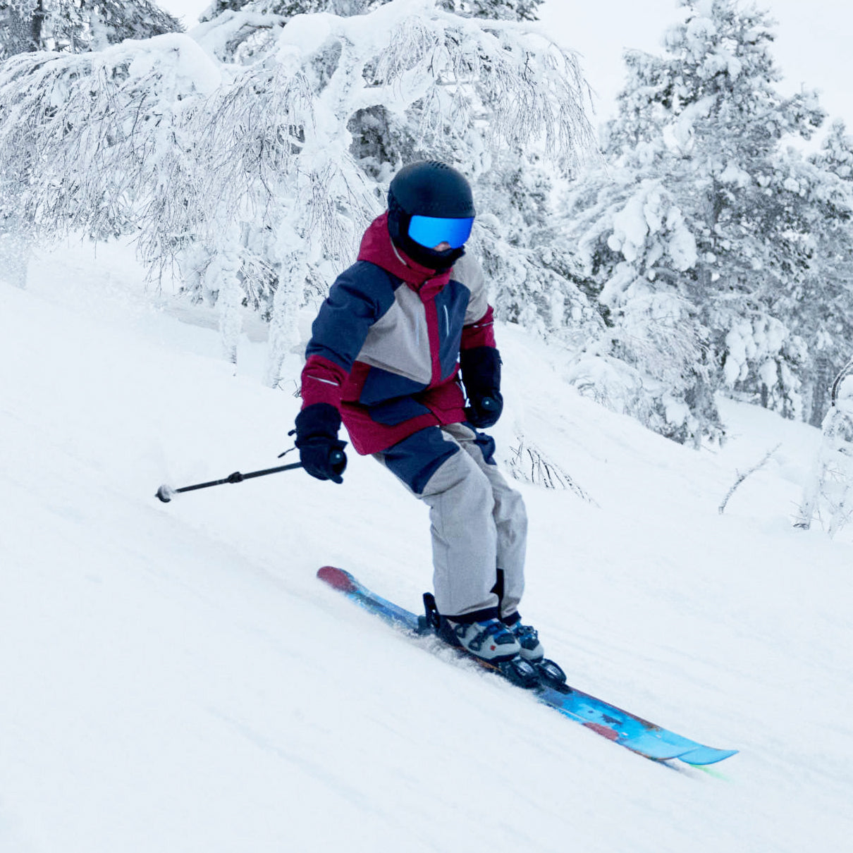 A skier in a red, navy, and gray winter jacket, light gray pants, and a black helmet with blue-tinted goggles is carving through deep snow on a snowy mountain slope with snow-covered trees in the background.