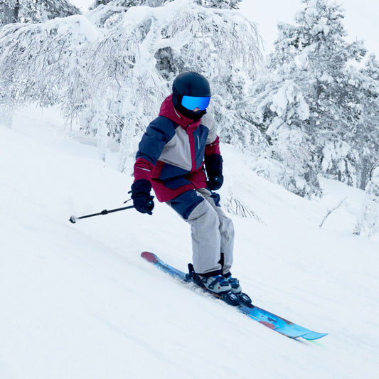 A skier in a red, navy, and gray winter jacket, light gray pants, and a black helmet with blue-tinted goggles is carving through deep snow on a snowy mountain slope with snow-covered trees in the background.