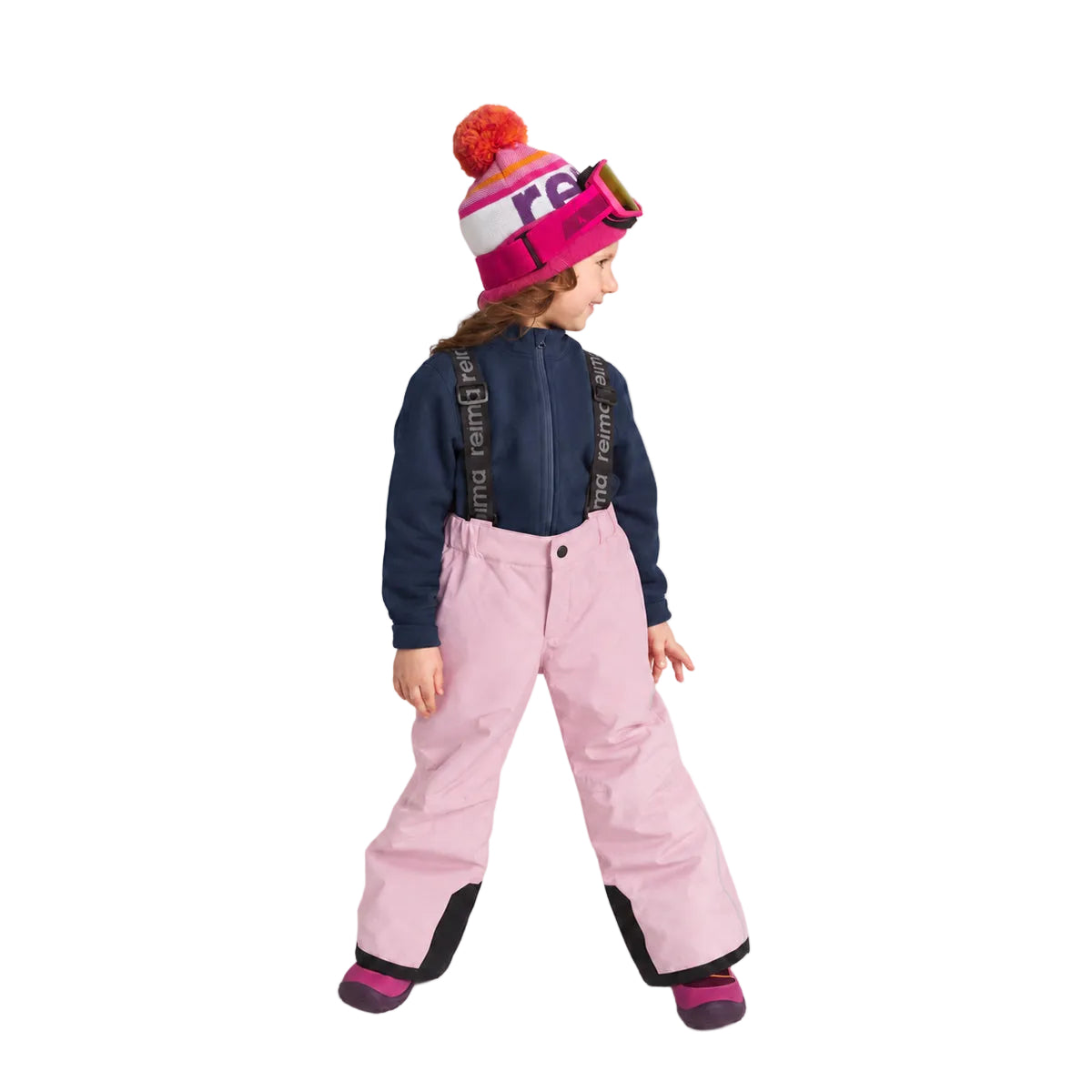A child wearing a pink ski jacket, light pink snow pants, a navy blue zip-up top, and a pink and white beanie with a red pom-pom, standing against a light gray studio background.