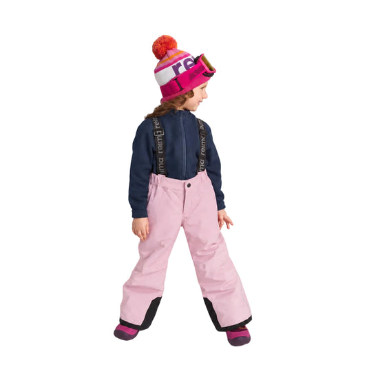 A child wearing a pink ski jacket, light pink snow pants, a navy blue zip-up top, and a pink and white beanie with a red pom-pom, standing against a light gray studio background.
