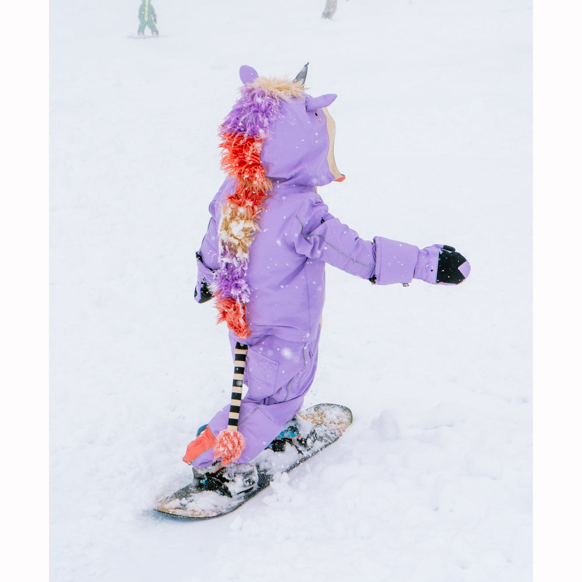 Child in a purple unicorn snowsuit snowboarding on a snowy background