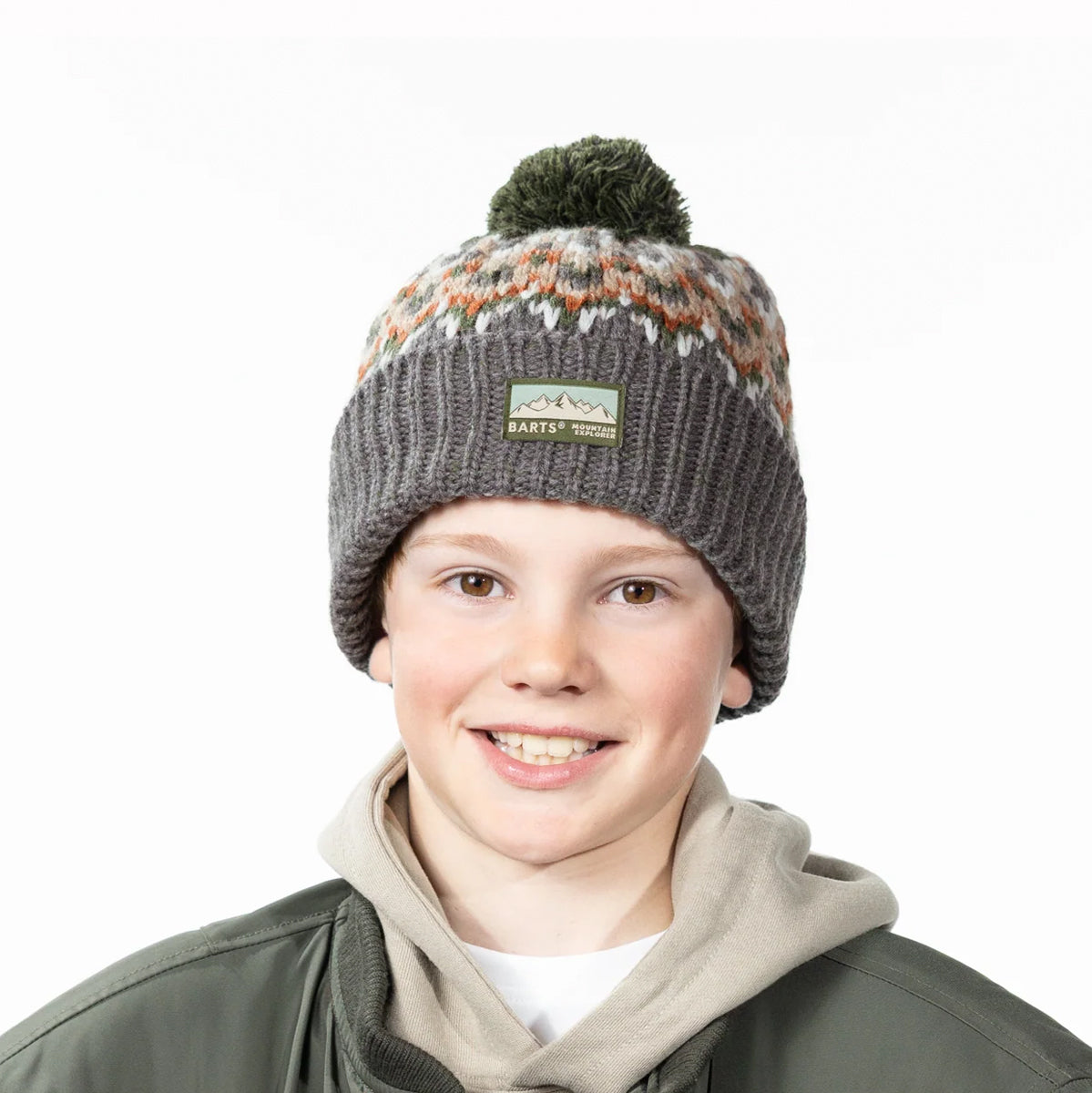 A smiling young person wearing a knit pom-pom beanie in dark gray with a multicolor pattern near the top and a visible Harter's logo patch. The background is a clean white studio backdrop.