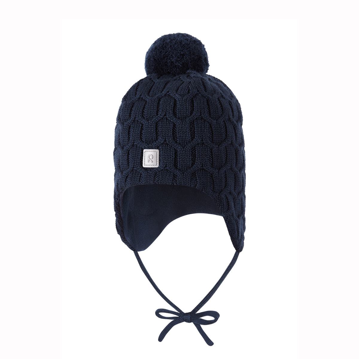 A navy blue cable-knit pom-pom beanie with ear flaps and chin ties, featuring a small square metal brand plaque on the front.