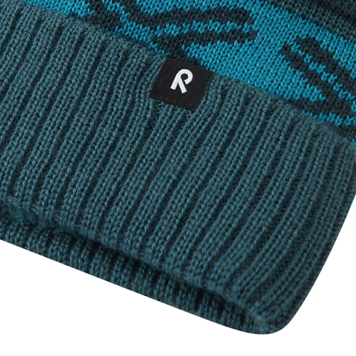 Close-up of a ribbed knit beanie in dark teal with a turquoise patterned section and a small black brand tab featuring a white stylized letter R.