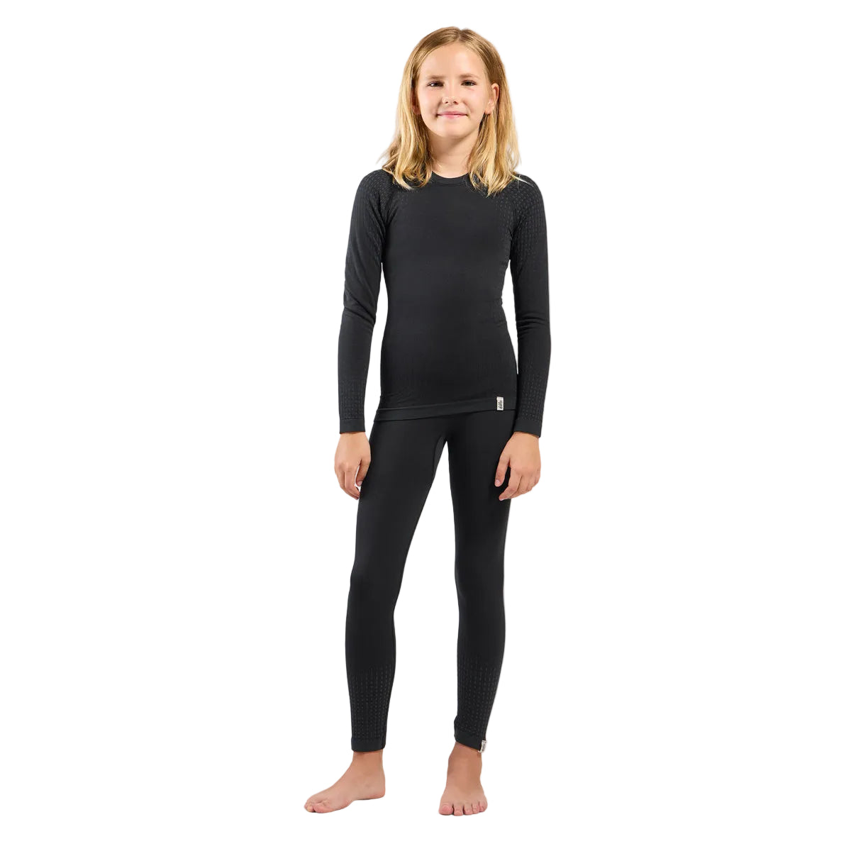 A young girl with blonde hair stands barefoot against a light gray studio background, wearing a black long-sleeve crewneck top and matching black full-length leggings.