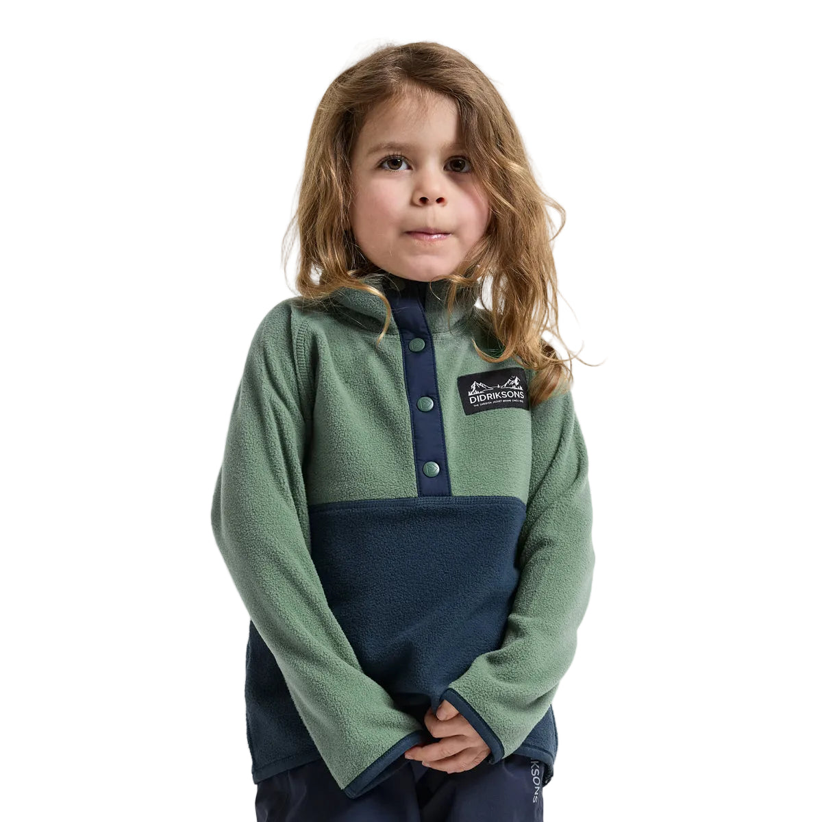 A young girl with long light brown hair wearing a color-block fleece pullover. The top features a sage green upper section and a dark navy blue lower section, with a matching navy button placket and a small black brand patch on the chest.